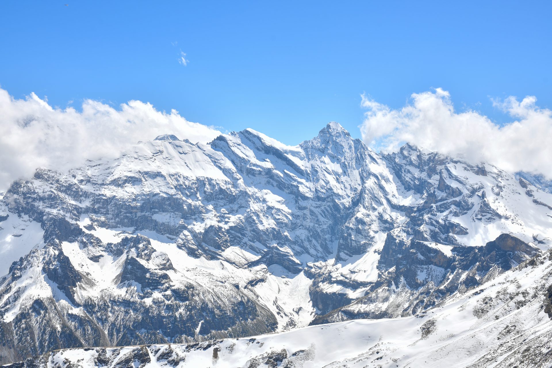 Snow-covered mountain peaks beneath a blue sky, with clouds along the ridge.
